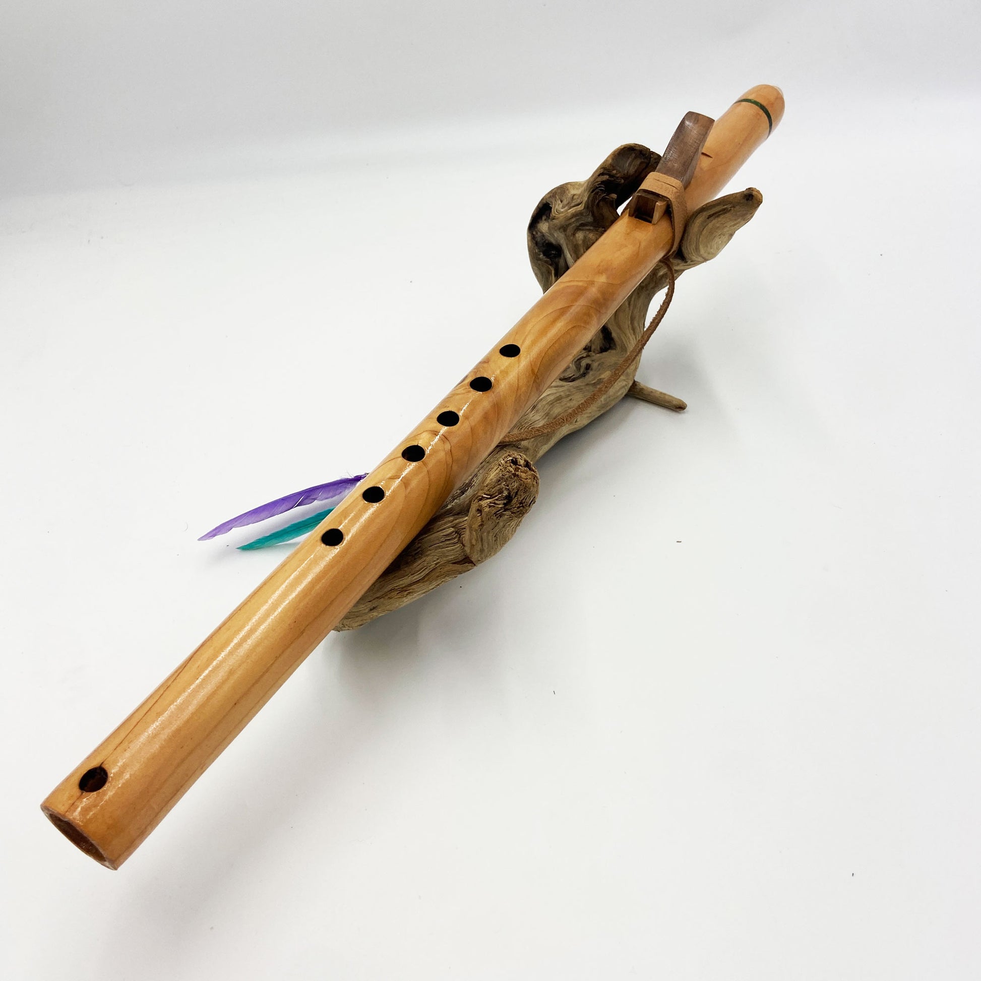 Flûte amérindienne artisanale en bois de cèdre clair, totem en noyer, ornements plumes violettes et turquoises, posée sur un support en bois flotté naturel.