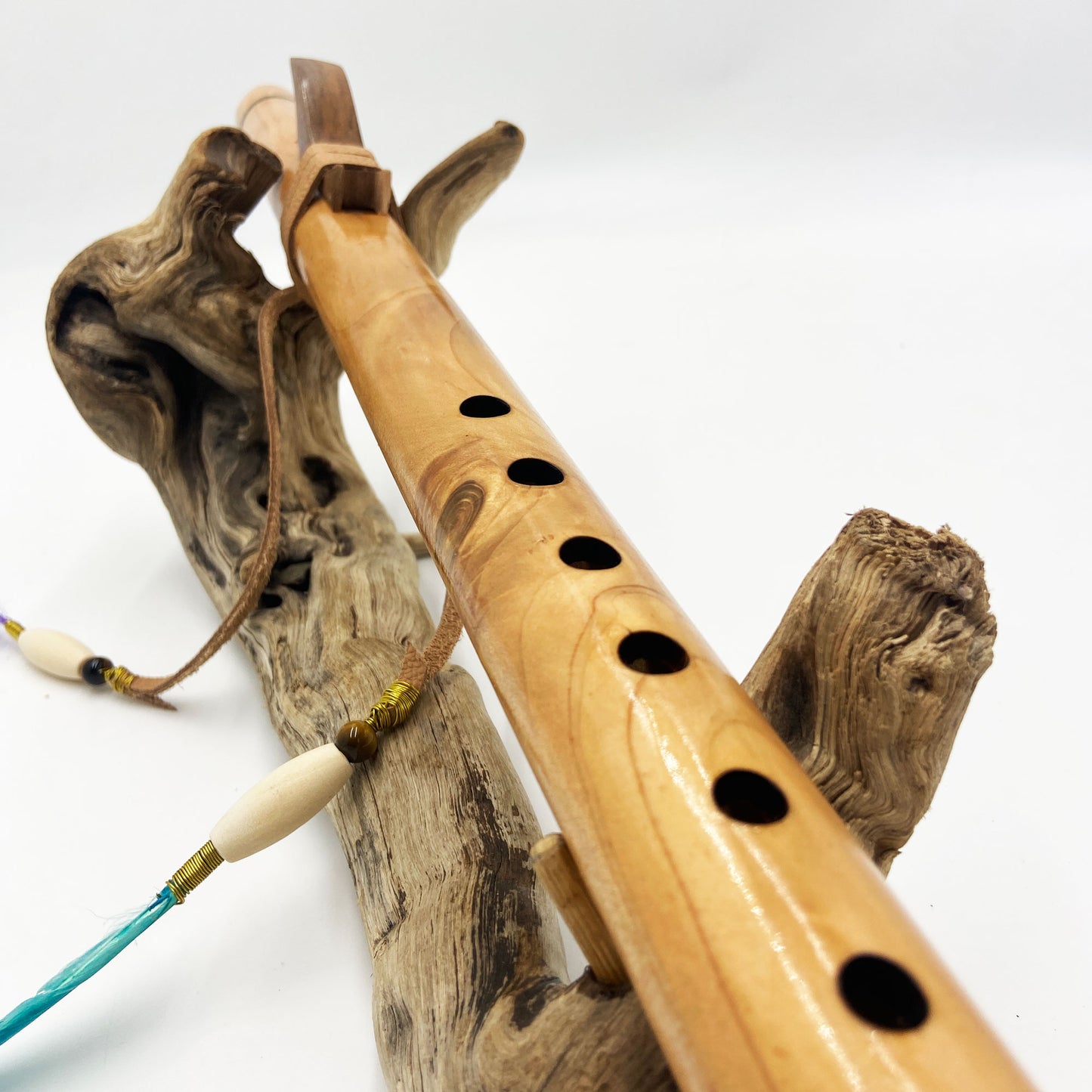 Flûte amérindienne artisanale en bois de cèdre clair, totem en noyer, ornements plumes violettes et turquoises, posée sur un support en bois flotté naturel.