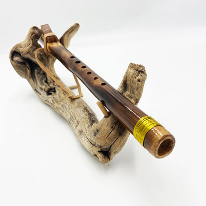 Flûte amérindienne en bambou effet caramel et noirci, embouchure en bois d'olivier veiné, totem en noyer et lacet de cuir sur support en bois flotté. Détail de fil doré à la base.