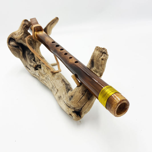 Flûte amérindienne en bambou effet caramel et noirci, embouchure en bois d'olivier veiné, totem en noyer et lacet de cuir sur support en bois flotté. Détail de fil doré à la base.