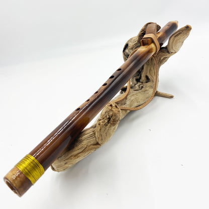 Flûte amérindienne en bambou effet caramel et noirci, embouchure en bois d'olivier veiné, totem en noyer et lacet de cuir sur support en bois flotté. Détail de fil doré à la base.