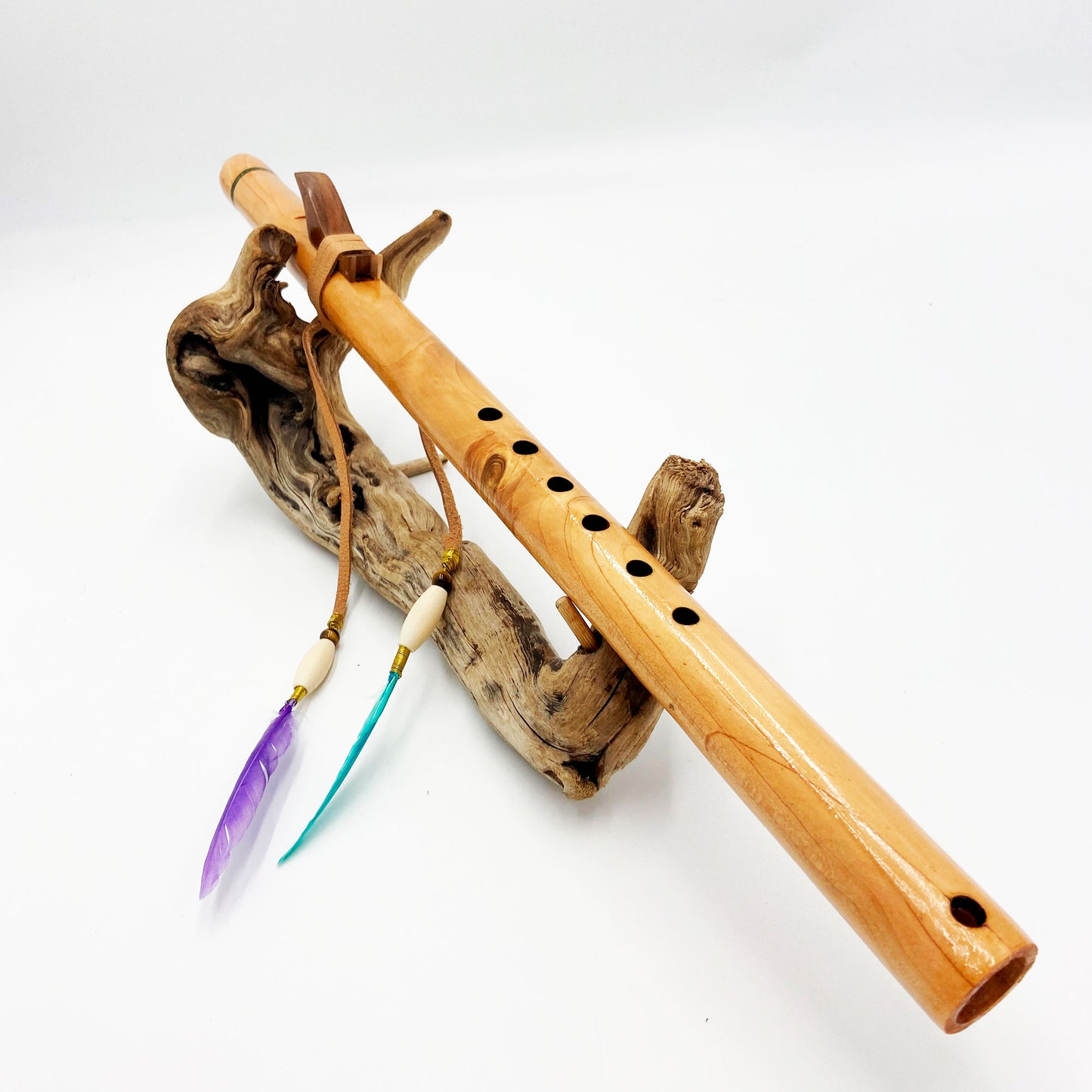 Flûte amérindienne artisanale en bois de cèdre clair, totem en noyer, ornements plumes violettes et turquoises, posée sur un support en bois flotté naturel.