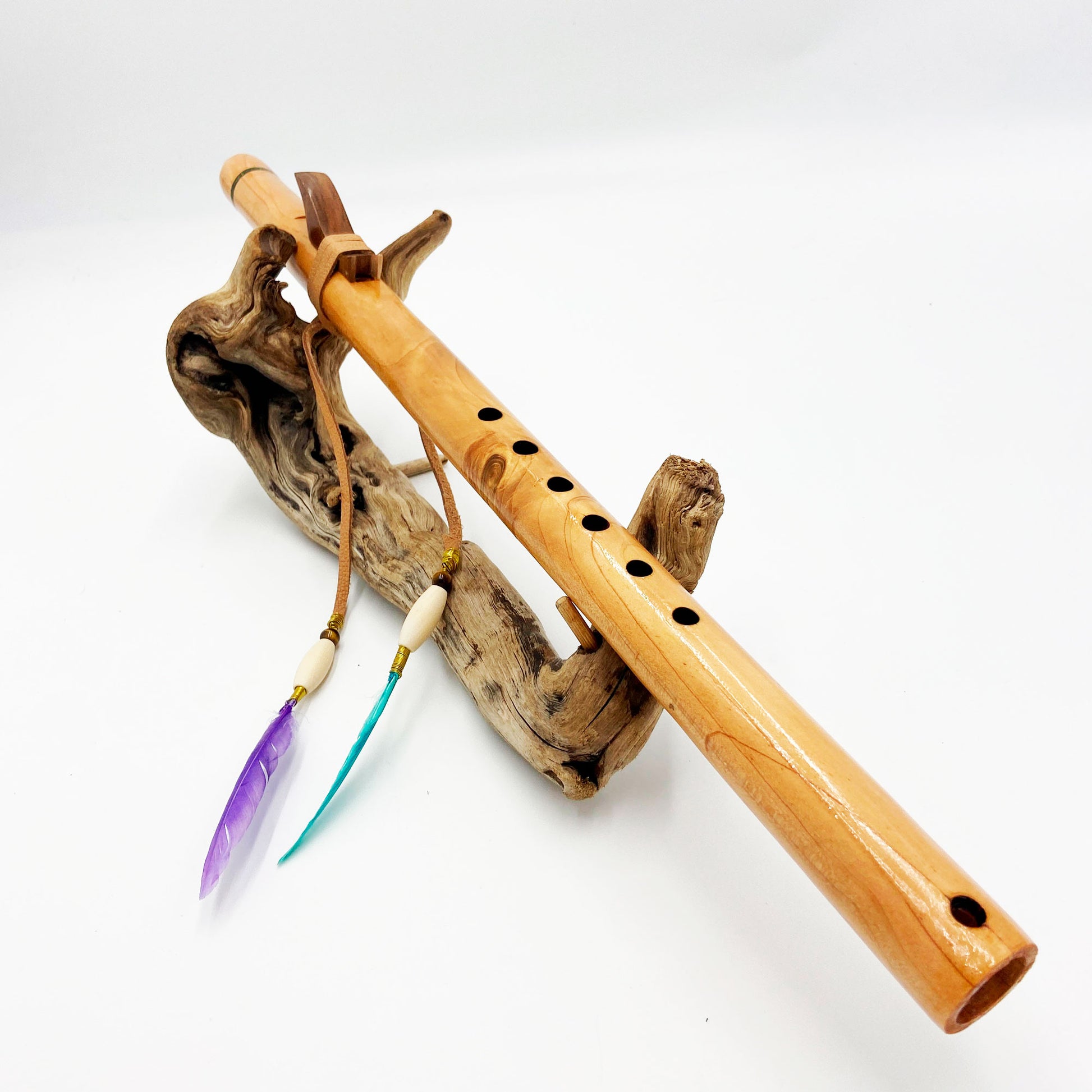 Flûte amérindienne artisanale en bois de cèdre clair, totem en noyer, ornements plumes violettes et turquoises, posée sur un support en bois flotté naturel.